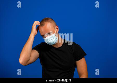 Portrait of young man with surgical medical mask in black casual style shirt, scratching his head, looking away and thinking what to do. indoor studio Stock Photo