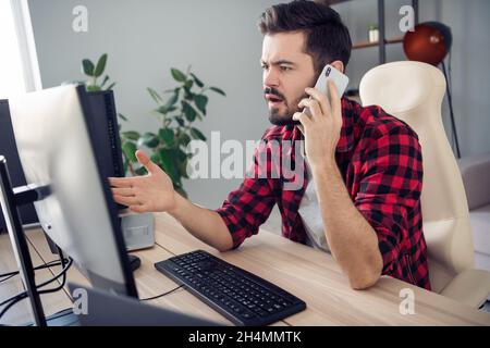 Photo of unhappy troubled young man hold phone talk editor look screen sit table inside office indoors workplace Stock Photo