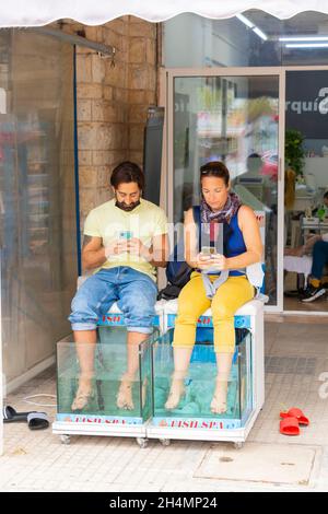 couple on their phones sit while having their feet nibbled by fish in Can Pastilla Majorca Spain Stock Photo