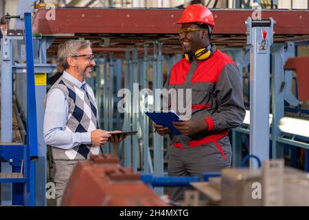 Smiling Multiracial Industrial Co-Workers Standing In A Factory And Talking Beside Next To Production Line. Stock Photo