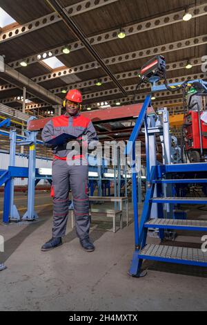 Full Figure Portrait Of Black Industrial Worker In Red Helmet, Yellow Safety Goggles And Work Uniform In A Factory. Stock Photo