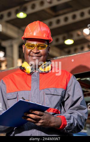 Waist Up Portrait Of Black Industrial Worker In Red Helmet, Yellow Safety Goggles And Work Uniform In A Factory. Stock Photo