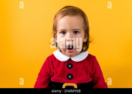 Portraite Emotion Cute Happy Cheerful Chubby Baby Girl in Santa Suit Yawns As If Singing At Orange Background. Child Christmas Scene Celebrating Stock Photo