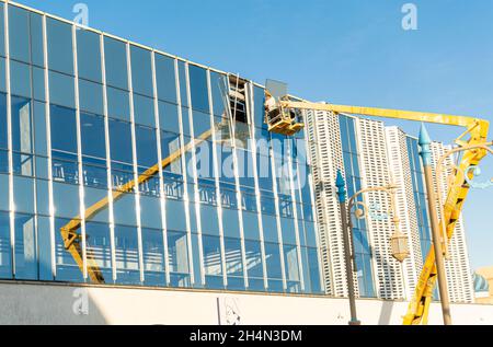 Workers on aerial platform instal glass piece on the facade of modern newly built glass office building in historic center of Turkistan, Kazakshtan Stock Photo