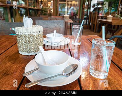 Thai restaurant setting table interior. Glasses of water with straw, plates and tissue paper on wooden table. Stock Photo