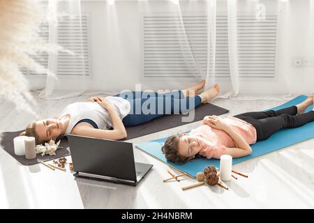 Two young beautiful sporty girls doing hatha yoga together in a modern light studio. They lie on the floor and meditate with their eyes. Yoga lessons Stock Photo