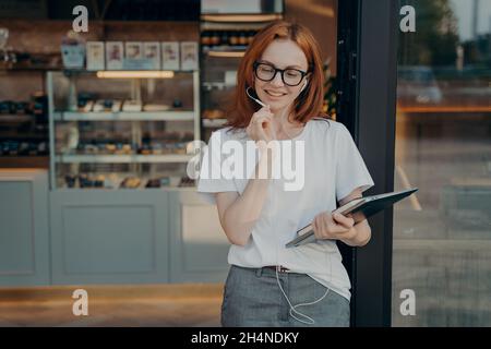 Positive redhead woman uses earphones with built in microphone tablet to communicate Stock Photo