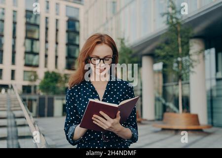 Outdoor shot of red haired woman makes notes in notepad makes plannings for working day Stock Photo
