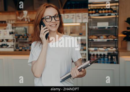 Happy beautiful businesswoman talking on cell phone outside, standing with laptop near coffee shop Stock Photo
