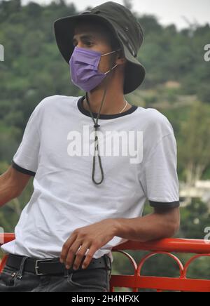 A Indian young man standing outside with wearing boonie hat, white t-shirt and face mask during coronavirus pandemic with looking at camera Stock Photo