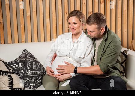 Man hugs pregnant wife sitting on cosy couch in living room Stock Photo