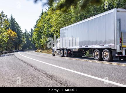 Classic big rig day cab white bonnet semi truck transporting commercial cargo in dry van semi trailer running for delivery on the winding highway road Stock Photo