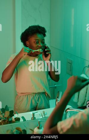 African young woman standing in front of the mirror and brushing her teeth while talking on mobile phone in the bathroom Stock Photo
