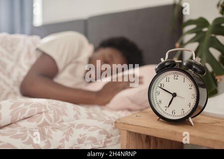 Close-up of alarm clock standing on the bed table with woman sleeping in her bed in the background Stock Photo