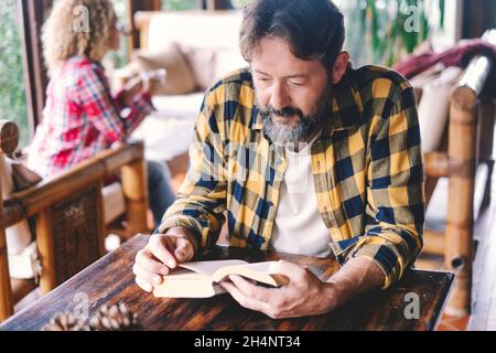 Adult man reading a book at home sitting on the wooden table with wife woman in background. Happy caucasian couple of people enjoying leisure time ind Stock Photo