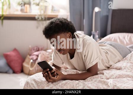 African young woman lying on the bed and watching something on her mobile phone in the room Stock Photo