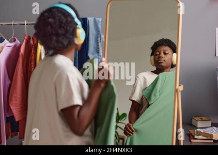 African woman in wireless headphones trying on the new dress while standing in front of the mirror in the room Stock Photo