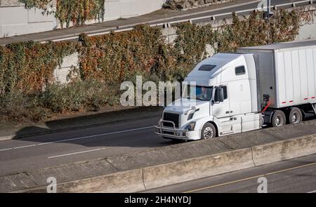 Classic big rig white bonnet semi truck with high roof transporting commercial cargo in dry van semi trailer running for delivery on the highway with Stock Photo