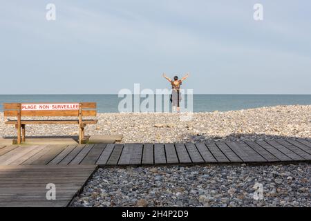 Lady alone facing the sea, making arm movements, on a pebble beach in Cayeux-sur-Mer. Opal Coast, France Stock Photo