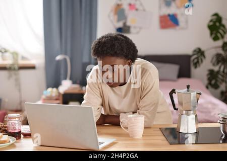 African young woman working online on laptop while sitting at the table and having breakfast at home Stock Photo