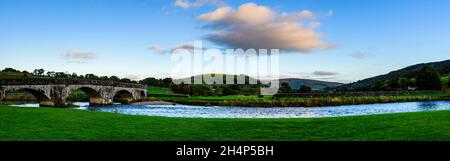 Panoramic evening riverside view of historic stone arched road bridge spanning flowing water of River Wharfe - Burnsall, Yorkshire Dales, England, UK. Stock Photo