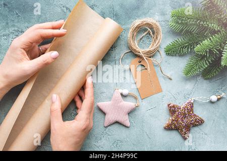 Close-up of hands holding a roll of wrapping craft paper on textured background with Christmas toys, tag and spruce branch. Cozy Christmas concept Stock Photo