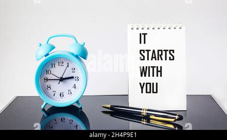 It all starts with you, text on a notebook, next to an alarm clock and a pen on a glass black background Stock Photo