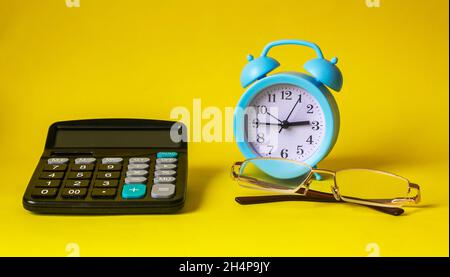 On a yellow background lies a calculator, glasses and a blue alarm clock Stock Photo