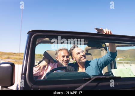 Smiling caucasian gay male couple taking selfies sitting in car at seaside Stock Photo