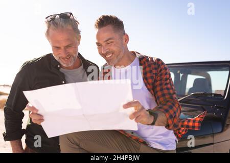 Happy caucasian gay male couple reading map, smiling by a car at seaside Stock Photo