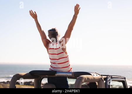 Rear view of caucasian man raising arms sitting on car on sunny day at the seaside Stock Photo