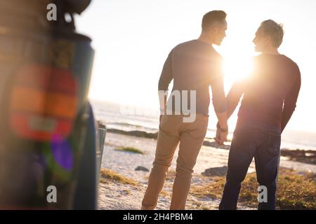 Happy caucasian gay male couple holding hands, walking away from car at seaside Stock Photo