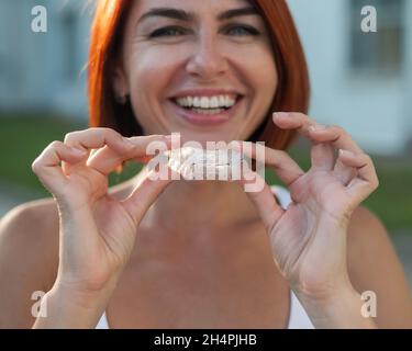 Red-haired Caucasian woman holding transparent mouthguards for bite correction outdoors. A girl with a beautiful snow-white smile uses silicone braces Stock Photo
