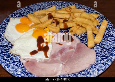 Ham eggs and chips on a round blue and white flower patterned porcelane plate on a dark wood table. Double yolk and single yolk eggs with brown sauce Stock Photo