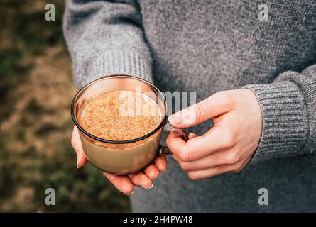 Close up view of woman hands wearing gray knitter cozy sweater holding cup of warm Chai latte with cinnamon powder outdoors in autumn. Stock Photo
