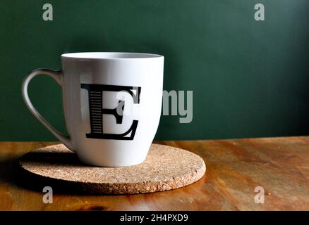 Large white mug with the letter 'E' on a cork mat on a wooden table set against a green background Stock Photo