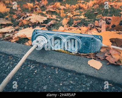 The hub of electrical outlets with the electric fork and cord on is on the ground in the autumn park among the leaves. Stock Photo