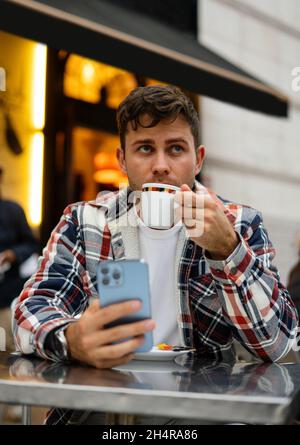 Handsome male in stylish outfit using smartphone while enjoying refreshing coffee and sitting at table in outdoors cafe while looking away Stock Photo