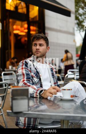 Pensive male in casual clothes sitting at table with mug of refreshing drink and enjoying coffee break in outdoors cafe Stock Photo