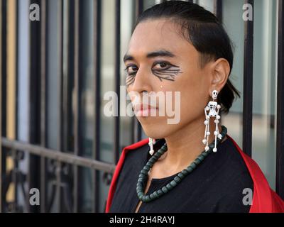 Handsome young non-binary transgender Mexican model with Day of the Dead skeleton earrings looks at the viewer in front of black burglar bars. Stock Photo