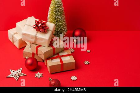 A stack of gifts in kraft paper and kraft boxes on a red background with wooden eco decorations for the Christmas tree. Christmas background, boxing d Stock Photo