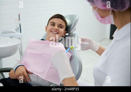 Handsome boy sitting in dentist chair smiles with beautiful toothy smile looking at a pediatric dentist teaching him about oral hygiene during dental Stock Photo