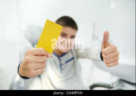 Soft focus on blank yellow visit card with copy space for insert ads and text in the hand of blurred handsome boy smiling toothy smile and showing thu Stock Photo