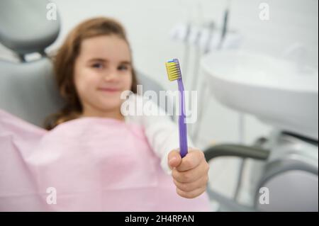 Focus on a toothbrush in the hand of adorable beautiful girl cutely smiling looking at camera sitting on dentist's chair during dental check-up. Oral Stock Photo