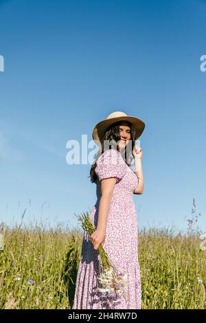 Portrait of beautiful young woman standing in field or meadow and holding bouquet of wildflowers Stock Photo