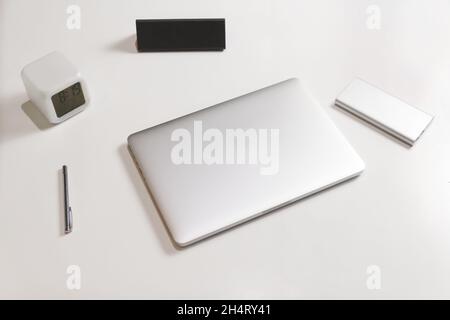 Side view of business working place with closed silver laptop, pen, clock, case for eyeglasses, power bank. Workspace on white table. White minimalist Stock Photo