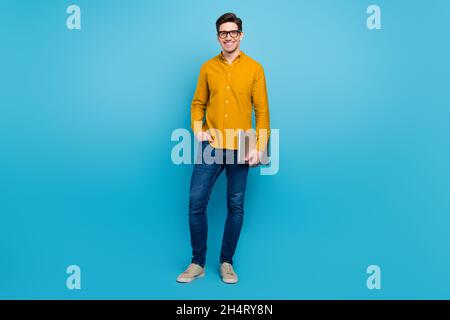 Photo of cheerful freelancer programmer guy hold pc wear yellow shirt jeans shoes isolated blue color background Stock Photo