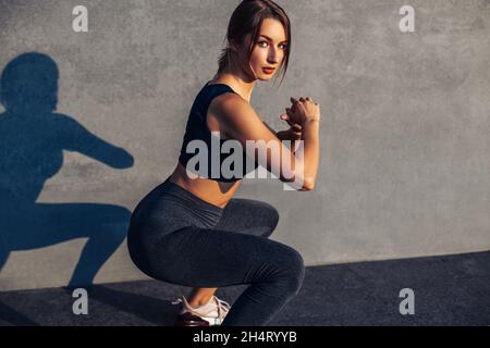 Image of a sportive woman, in sneakers and a tracksuit squatting, doing squats on the street isolated against a gray city wall, fitness, sport, exerci Stock Photo