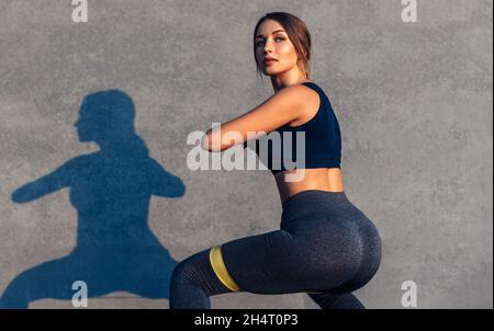 Fitness woman doing buttocks exercises with resistance band on gray street background, sportive woman working out doing squats, doing sports outdoors Stock Photo