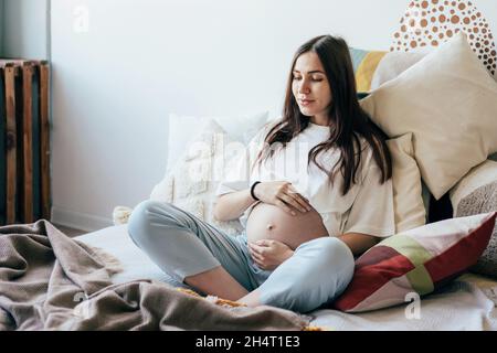 Young caucasian pregnant pretty brunette woman is sitting on the bed in the apartment and touching her belly in the ninth month of pregnancy. Waiting Stock Photo
